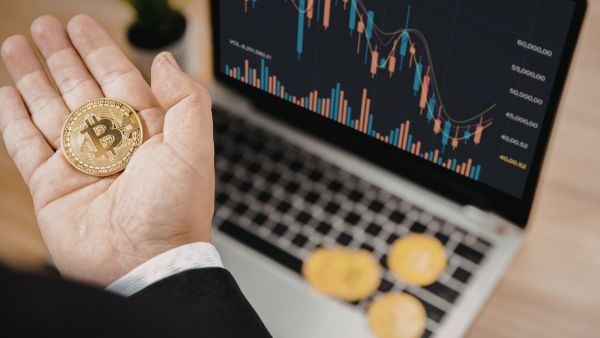 Investidor segurando moeda de bitcoin próximo a computador com informaçoes sobre criptoativos